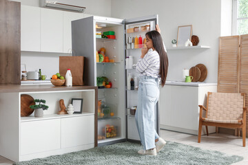 Thoughtful young woman near open fridge in kitchen
