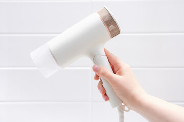 Female hand holding hair dryer against white tiles background in bathroom close-up.