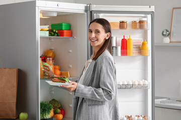 Young businesswoman taking sandwich and juice from open fridge in kitchen