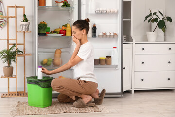 Young woman throwing spoiled food from open fridge in kitchen