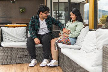 Husband with prosthetic leg touching pregnant wife's belly on patio sofa