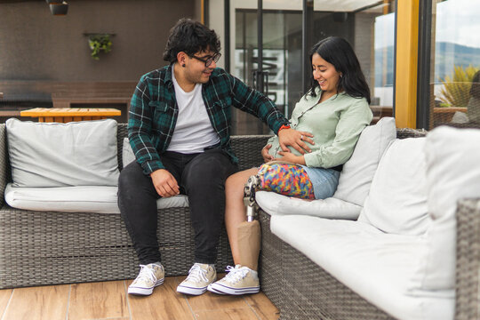 Husband with prosthetic leg touching pregnant wife's belly on patio sofa - Powered by Adobe