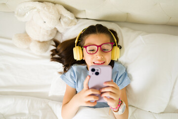 Smiling girl wearing headphones using smartphone in bed
