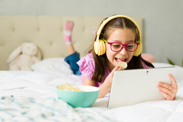 Preteen girl eating potato chips and watching tablet on bed