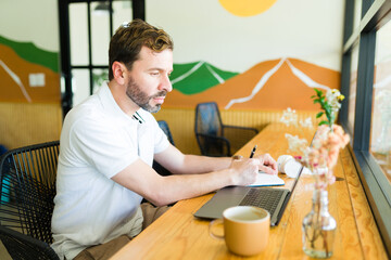 Focused man working remotely in cozy coffee shop, taking notes and using laptop