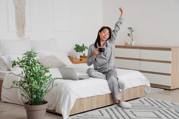 Young Asian woman with cup of coffee stretching in bedroom
