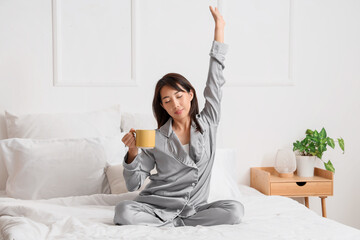 Young Asian woman with cup of coffee stretching in bedroom