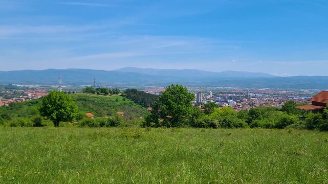 Sunny spring day in southern Serbia: rooftops of Leskovac peek through undulating green hills under a vast blue sky in this wide panoramic landscape