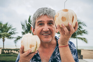 happy old man tourist holds a drinking coconut with a straw on vacation on a beach by sea in the...