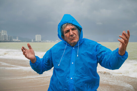 sad old man tourist in a raincoat on a beach by sea with waves in cloudy and rainy weather. Concept of the rainy and monsoon season