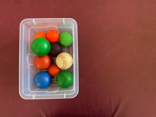 Vibrant collection of colorful stress balls and a baseball in a clear plastic container, set against a solid reddish-brown background, viewed from above.