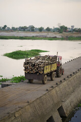 Tractor transporting logs on river bridge, Rural tractor with timber load near river, Wood...