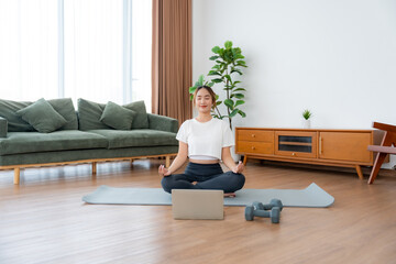 Young Asian woman meditating in lotus position at home, sitting on the floor in fitness clothing....