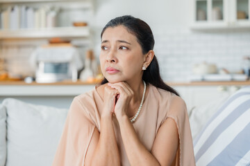 Upset Asian senior woman sitting on sofa, lost in thoughts, feeling lonely, disappointed, alone at...