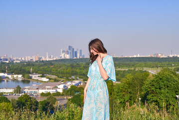 A woman in a navy blue dress is standing on a hill with a panoramic view of the city below. The sky...