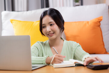 Young asian woman planning and strategy about finance and investment or tax while writing on textbook in living room at home, woman with expense and budget of home, business and finance.