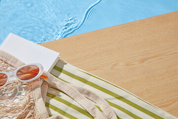 Summer poolside scene with striped towel, sunglasses, woven bag, and notebook on wooden deck near clear blue water, perfect for vacation and product mockups.