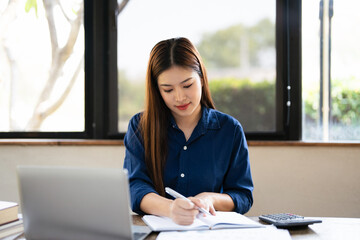  Focused Asian business woman sits in home office taking notes in a notebook, working on her...