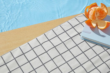 Poolside flat lay with peeled orange, sunglasses, and book on checkered towel by wooden deck and...