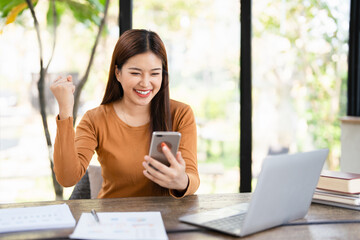  Overjoyed woman looking at phone screen, celebrating success, showing yes gesture, sitting at work...