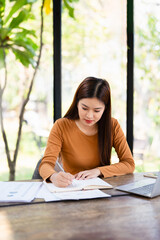 Focused Asian business woman sits in home office taking notes in a notebook, working on her...
