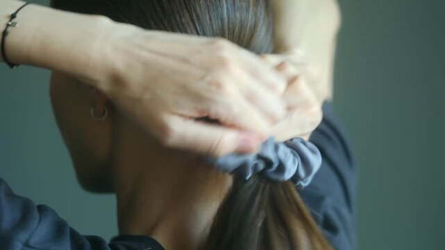 Styling long brown hair into elegant ponytail using gray scrunchie, demonstrating simple hairstyling technique with precise step by step movements. Woman making a ponytail with scrunchie