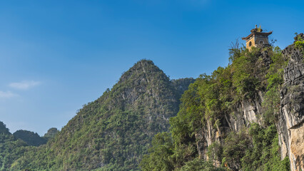 Fototapeta premium Steep mountains against the blue sky. Green forest on the slopes. At the top, you can see a Chinese pagoda with a roof with curved cornice edges. China. Guilin