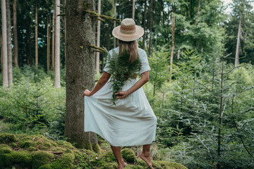 Serenity in the woods as a woman in a summer dress and hat stands facing away, enjoying the...