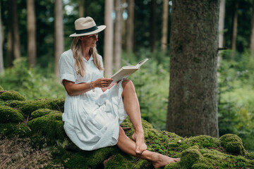 White dress woman relaxes on moss in forest, reading a book under green trees