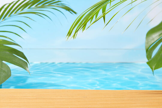 Tropical infinity pool with wooden deck, palm leaves, and bright blue water under clear sky, offering a serene and modern background for vacation or product visuals. - Powered by Adobe