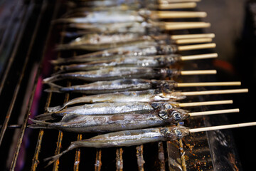 Delicious grilled Shishamo (Japanese smelt or capelin), seasoned with salt and filled with roe, being cooked on a hot barbecue grill. A popular and savory Japanese street food or snack.
