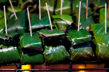 Traditional Northern Thai Jîn Som Mok Yang (fermented pork wrapped in banana leaves) being grilled. A popular savory local delicacy and street food, showcasing authentic Thai cuisine.
