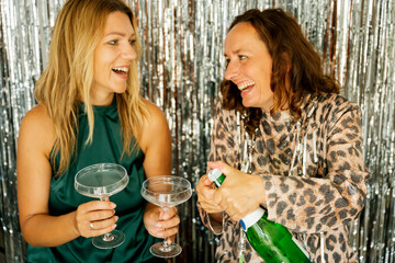 Two happy women opening sparkling wine bottle celebrating new year's eve