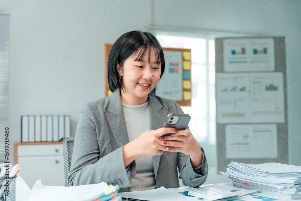 Poster young office worker using mobile phone while working at desk, she's smiling and looking at the scree