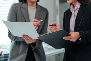 Two businesswomen collaborating in an office, holding pens while analyzing documents and reviewing...