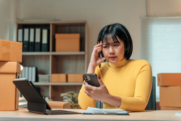 Young business woman holding head and looking worried while using mobile phone. Sitting at desk with cardboard boxes and digital tablet in modern office. Experiencing difficulties with online orders