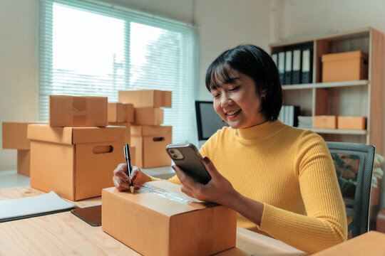 Young Asian woman business owner working in home office, writing customer addresses on cardboard boxes, preparing parcels for delivery while checking orders on smartphone