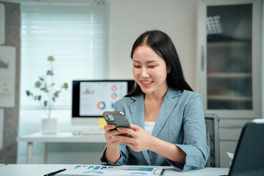 Asian businesswoman using smartphone and smiling while working with documents and computer at office desk, concept of modern technology and business administration