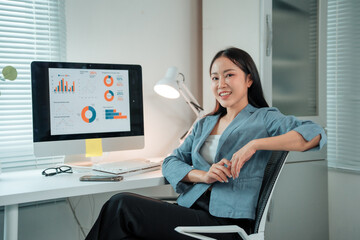 Young Asian businesswoman sitting in a chair in her office, smiling while working with charts and graphs displayed on her computer screen, demonstrating confidence and expertise in data analysis