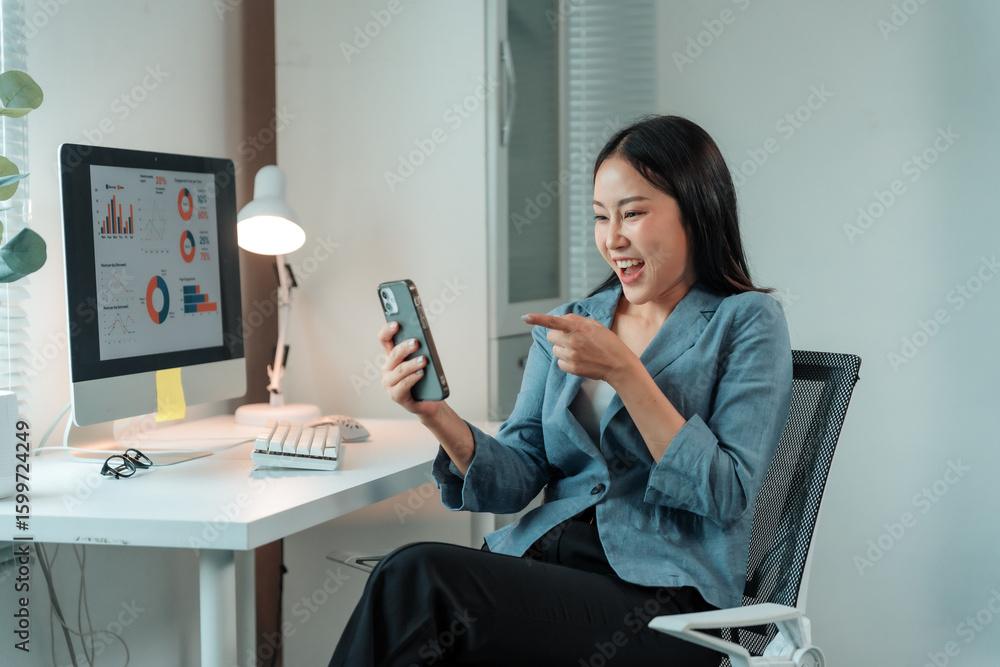 Poster excited businesswoman pointing at smartphone and celebrating success while sitting at desk and worki
