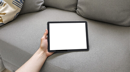 A hand holds a tablet with a blank white screen, resting on a gray fabric couch with pillows.  The scene is simple and domestic, suggesting relaxation or leisure