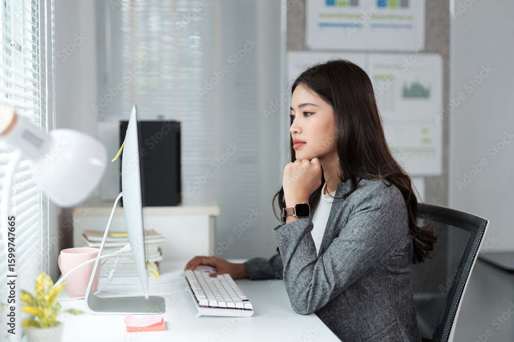 Poster young businesswoman wearing a gray suit and smartwatch is working with desktop computer in a modern 
