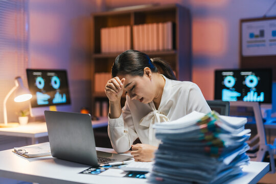 Tired businesswoman suffering from headache while working on laptop late at night in office with a big pile of paperwork and graphs on the table