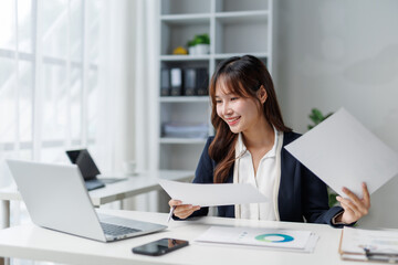 Asian businesswoman comparing documents while working on a laptop in a modern office, smiling...