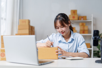 Young Asian woman entrepreneur is writing on a shipping label while using a laptop and smartphone...