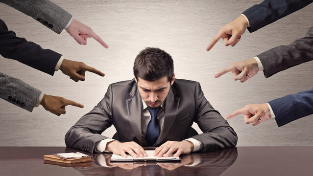 Pointing Fingers: A man in a suit sits with his head bowed, surrounded by hands pointing at him, conveying blame and pressure in a workplace environment.
