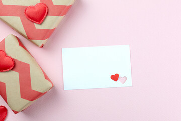 A blank white card with two delicate heart cutouts (one red, one pink) offers ample copy space for your text and gift box, red heart on pink background. Top view, flat lay.