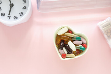 Light green heart shaped bowl with various colored dietary supplement pills and clock on pink...