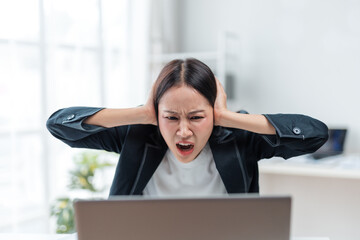 Young Asian manager is working in office, she is covering her ears with her hands and screaming,...