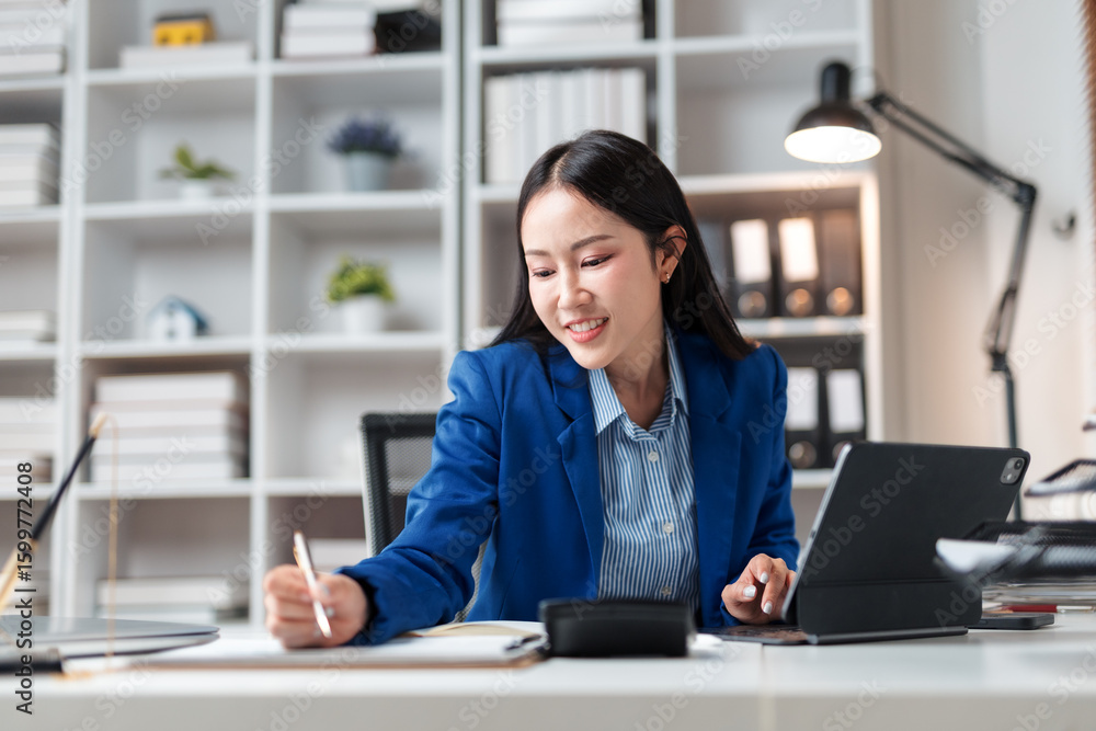 Poster young manager smiling and working with digital tablet and taking notes in her notebook, sitting at d